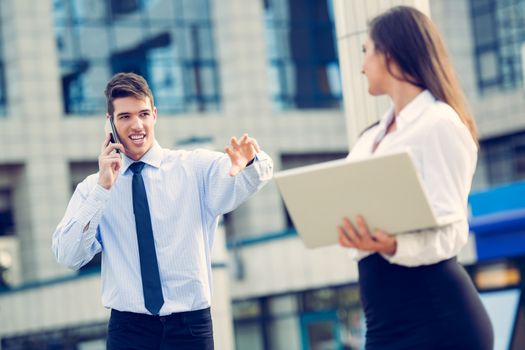 Young business partners standing in front of the building. Businessman talkin on a mobile phone while his woman partner  working on a laptop.