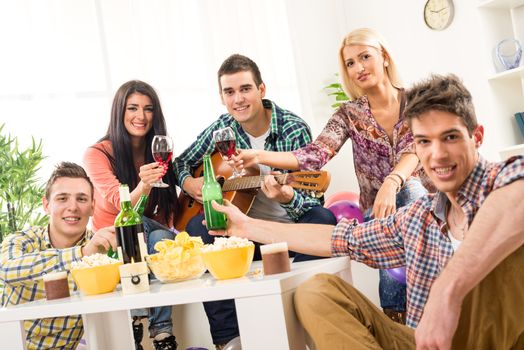 Small group of young people at the home party, enjoy the sounds of the acoustic guitar, drinking wine and beer. Looking at camera.