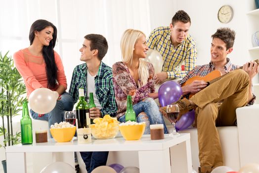 A small group of young people hang out at the house party, chatting with each other while their friend having fun playing acoustic guitar.