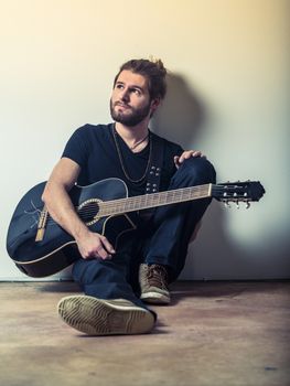 Photo of a young attractive man with long hair and beard sitting on the floor and holding an acoustic guitar. Filtered to look vintage.