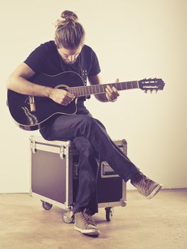 Photo of a young attractive man with long hair and beard sitting on a flight case and playing an acoustic guitar. Filtered to look vintage.
