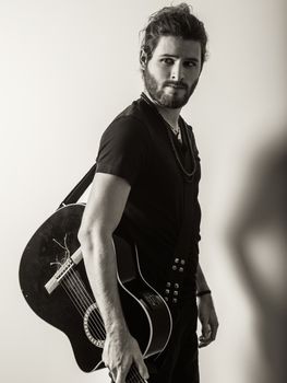 Photo of a young attractive man with long hair and beard standing and holding an acoustic guitar. Filtered to look vintage.
