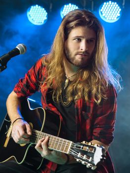 Photo of a young man with long hair and a beard playing an acoustic guitar on stage with lights and concert atmosphere.