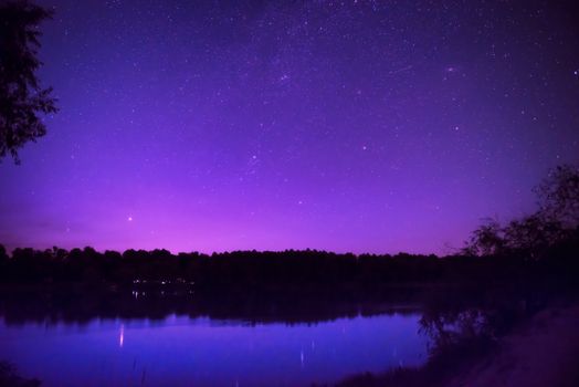 Beautiful purple night sky with many stars on a lake with forest on the other coast. Milkyway reflection in water