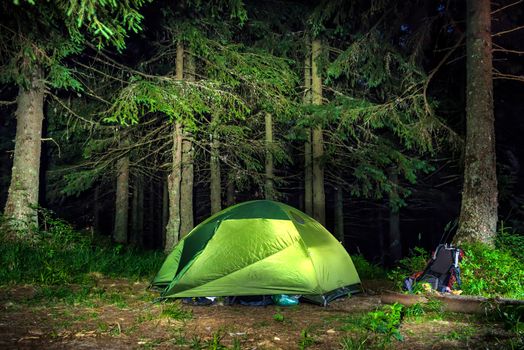 Camping in the forest. Green illuminated tent under dark night trees
