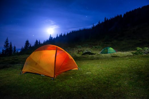 Two Illuminated orange and green camping tents under moon, stars at night