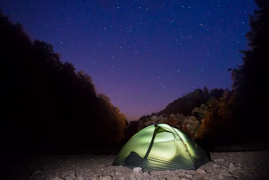 Illuminated green tent at night in the forest under dark blue sky with many stars