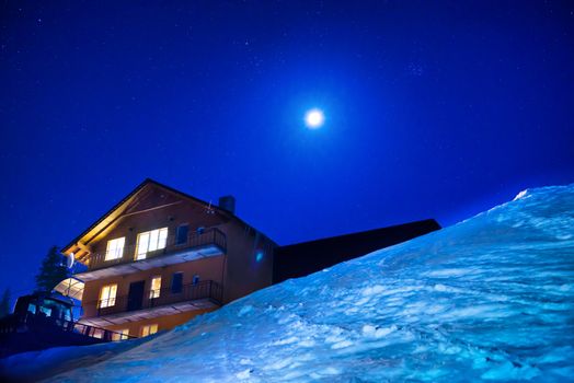 Christmas house in winter mountains at night. Snow landscape with blue dark sky with milkyway and many stars