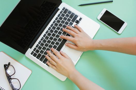 Hand using laptop on creative flat lay workspace desk with smartphone and stationery on green background