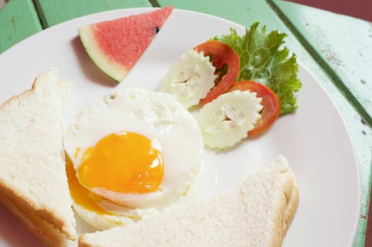 Close up fried egg, bread, cucumber, tomato and lettuce with watermelon in white plate on green wood table near window.