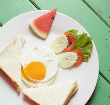 Fried egg, bread, cucumber, tomato and lettuce with watermelon in white plate on green wood table.