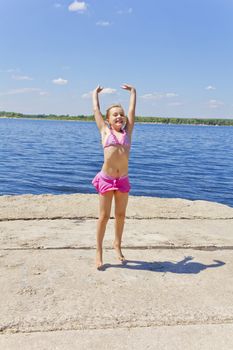 Cute jumping girl on the riverbank in pink swimsuit