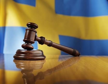 Mahogany wooden gavel on glossy wooden table, flag of Sweden in the background.
