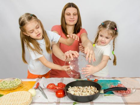 Mother with two daughters at the kitchen table with a plate of mushrooms is poured into the pan