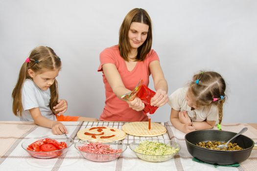 Two little girls enthusiastically watched as mum pours ketchup basis for pizza
