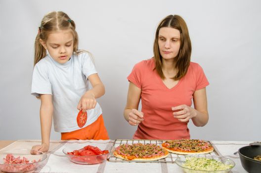 Six year old girl takes a plate of cutting tomatoes for pizza under the supervision of mum