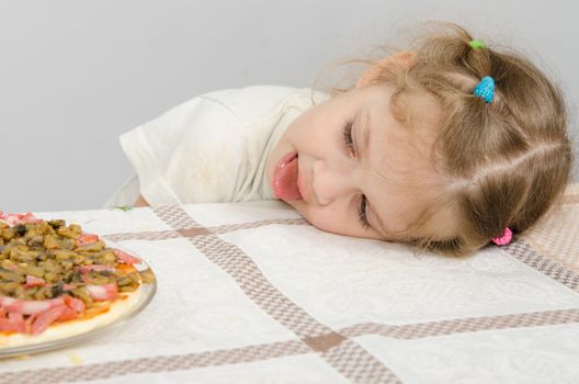 Little girl with protruding tongue rested her head on the table and looks at the pizza