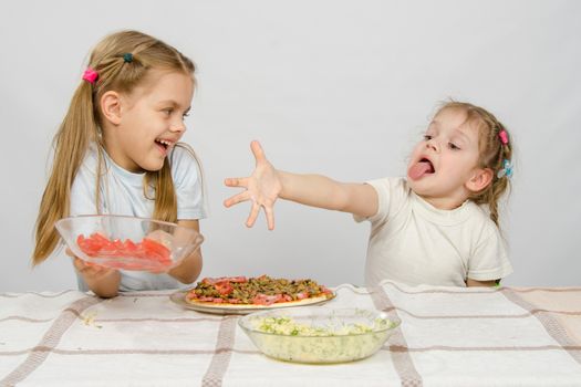 Two little girls at a table prepared pizza. One with a whimsical view stretches a hand to a plate with tomatoes, which takes the other with a smile