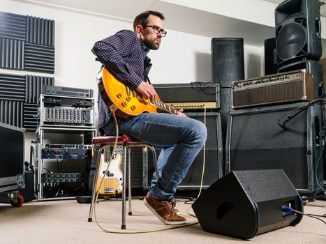 Photo of a man in his late 20's sitting in a recording studio playing his electric guitar.