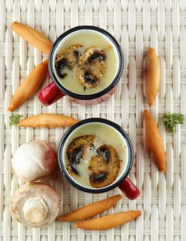 Delicious Homemade Mushrooms Cream Soup Decorated with Roasted Champignons in Two Red Cups with Bread Sticks closeup on Wicker background. Top View