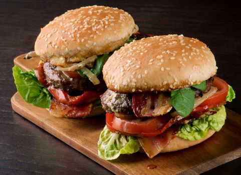 Two Tasty Hamburgers with Beef, Bacon, Lettuce, Tomatoes, Basil, Roasted Onion and Juicy Sauce on Sesame Buns In a Row closeup on Wooden Cutting Board