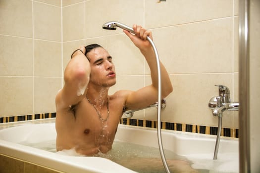 Handsome young man in bathtub at home having bath, washing body and hair with bathfoam and shampoo