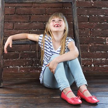 Girl 6 years old in jeans and a vest sits on the floor next to a brick wall and filled with laughter