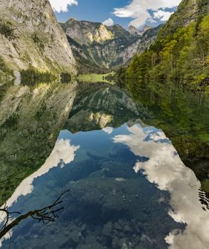 Reflection of blue sky and mountains in calm mountain lake with trees, impressive white clouds, with view on some stones under water near the shore and a valley in the background