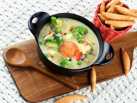 Delicious Homemade Vegetables Creamy Soup with Broccoli, Carrots, Zucchini, Leek, Red Bell Pepper and Green Pea in Black Iron Stewpot with Wooden Spoon and Bread Sticks closeup Wicker background
