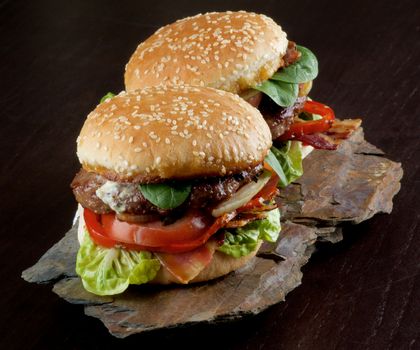 Two Tasty Hamburgers with Beef, Bacon, Lettuce, Tomatoes, Basil, Roasted Onion and Juicy Sauce on Sesame Buns on Stone Board closeup on Dark Wooden background