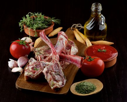Arrangement of Raw Lamb Ribs with Tomatoes, Garlic, Thyme, Rosemary, Spices and Olive Oil on Wooden Cutting Board closeup on Dark Wooden background