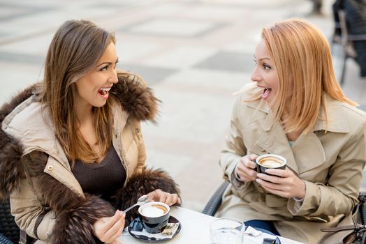 Young cheerful female friends sitting in a street cafe and enjoying coffee.