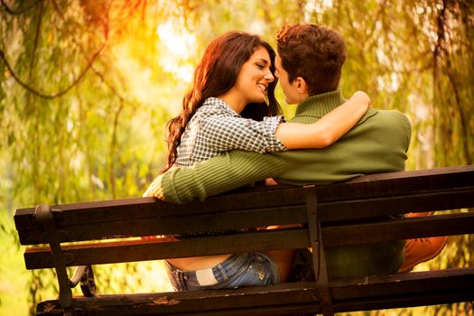 Rear view of a Young couple in love sitting on a park bench, illuminated by sunlight, passionate look at each other in the moment before the kiss.