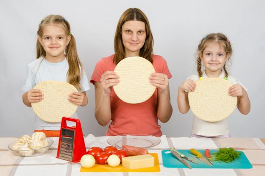 Mum with two little girls sitting in a row at the kitchen table and a hand-held pizza bases