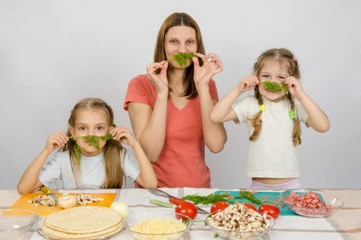 Young housewife with two daughters having fun holding sprig of parsley as a mustache at the kitchen table when sharing cooking