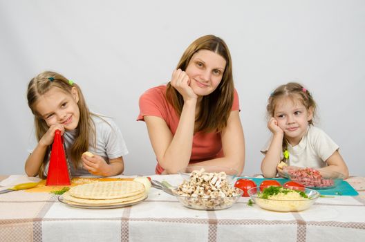 Satisfied mother with two daughters sitting resting his head in his hands at the table with the products for pizza
