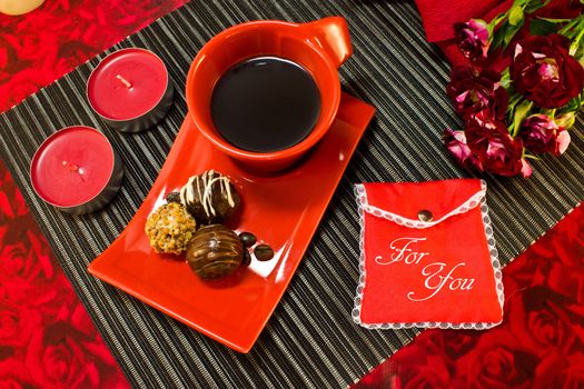 Coffee in red cup on a saucer with a chocolate, a bouquet of roses and gift envelope with the inscription for you