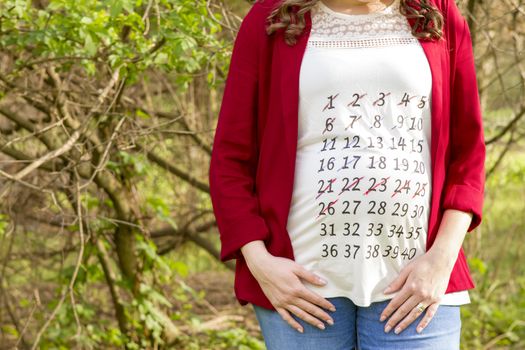 Pregnant woman in red jacket with calendar on her T-shirt outdoor in the park
