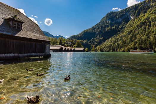 View from boats harbour Schönau over the Königssee in Berchtesgadener Land with returning ship, some brants in the water, boatshouses, Villa Beust in front of deep blue sky.