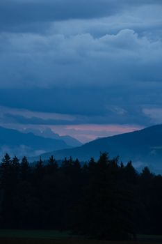 dark forest in front of a mountain chain with ascending mist, cloudy sky after sunset with reddish shining horizon