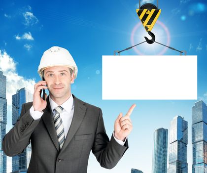 Smiling businessman in helmet talking on mobile with blank block on city background