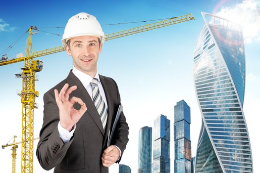 Smiling businessman in helmet holding papers and showing ok with building crane background