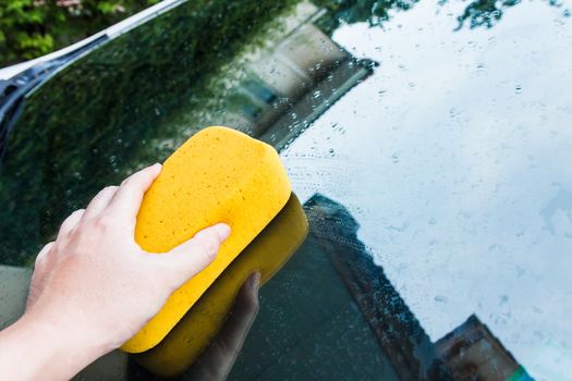 Hand cleaning wet car's window with yellow sponge.