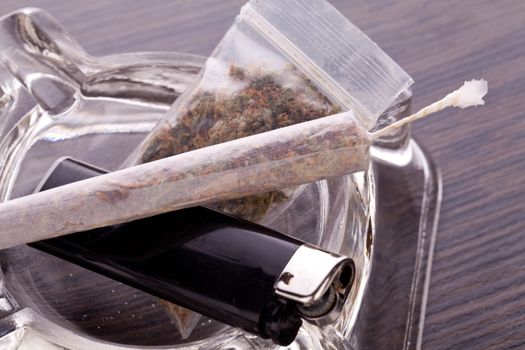 Close up of marijuana joint made with translucent rolling papers, plastic baggy of dried marijuana, black lighter and pipe on white background