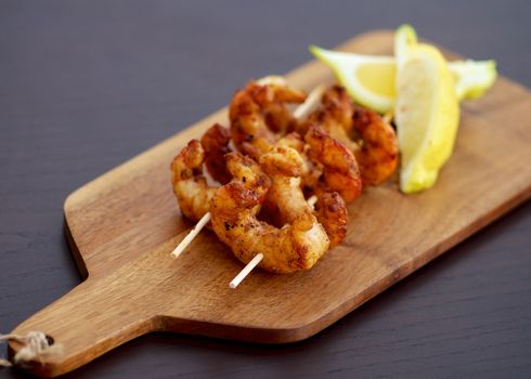 Delicious Grilled Prawns on Wooden Sticks with Sliced Lemon closeup on Wooden Cutting Board. Focus on Foreground