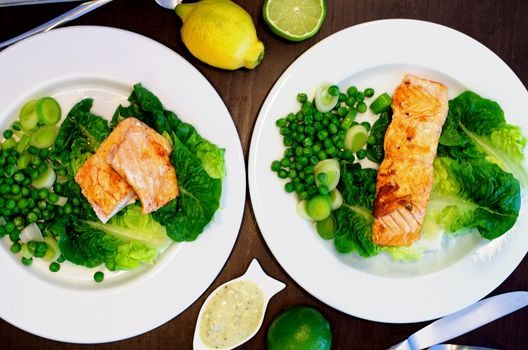 Delicious Roasted Salmon with Sweet Pea, Leek and Salad Romano on White Plates with Lemon and Sauce closeup on Dark Wooden background. Selective Focus