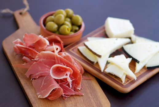 Traditional Spanish Tapas with Sliced Hamon, Goat Cheese and Green Olives on Wooden Cutting Board closeup on Dark Wooden background. Focus on Hamon
