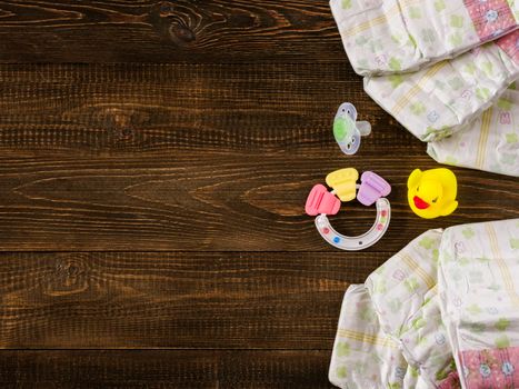 Japanese disposable diapers, teether, dummy and rubber duckling on dark wooden background with copy-space. Flat lay