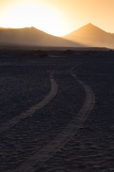 Car tire tracks in sand. Dirt road vanishing to the mountains sunlit in sunset. Copy space for text. Vertical composition.