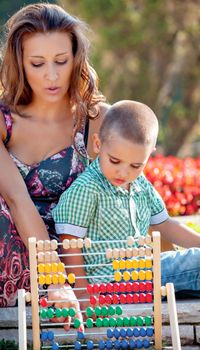 Mother teaches her son mathematics in the summer park on colorful wooden abacus.